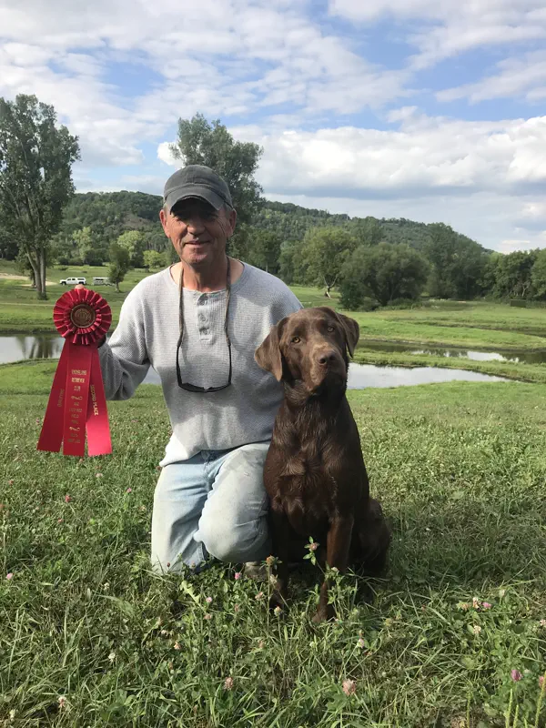 A handler kneeling in a grassy field with a chocolate Labrador Retriever and a red rosette