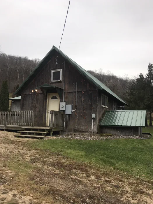 A rustic A-frame cabin with a green metal roof on a lawn backed by bare trees