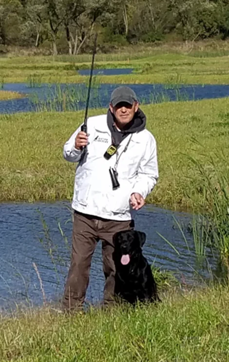 A man in a white jacket and gray cap stands in a grassy wetland holding a long rod, a black dog sitting beside him
