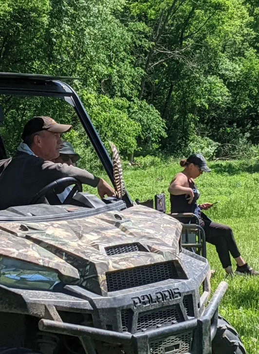 A camouflaged Polaris side-by-side parked in a grassy clearing with a driver aboard and a woman sitting on the grass nearby